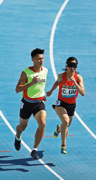 Imagem: Fotografia. Um homem com uniforme e colete ao lado de uma mulher de uniforme e uma venda nos olhos. Eles estão correndo em uma pista de corrida.  Fim da imagem.