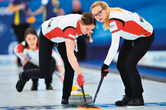 Imagem: Fotografia. Duas mulheres com uniforme, branco, laranja e preto em uma pista. Elas estão inclinadas segurando um objeto semelhante a uma vassoura próximo a um disco grande. Fim da imagem.