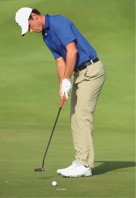 Imagem: Fotografia. Um homem com boné, camisa, calça, tênis e uma luva. Ele está segurando um bastão próximo a uma bola branca e um buraco.  Fim da imagem.