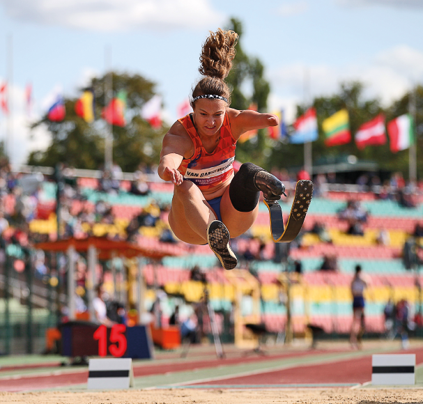 Imagem: Fotografia. Uma mulher com cabelos loiros, presos em um rabo de cavalo, usando prótese na perna esquerda. Ela está saltando sobre um campo de areia. Fim da imagem.