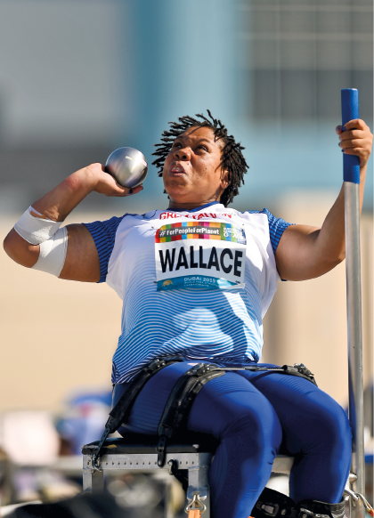 Imagem: Fotografia. Uma mulher robusta com cabelos trançados, sentada em um banco com cinto. Segurando com a mão esquerda um peso redondo e prateado.  Fim da imagem.