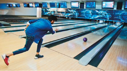 Imagem: Fotografia. Um homem usando calça e casaco jeans. Ele está inclinado em uma pista. Em frente, uma bola na pista em direção aos pinos. No fundo, televisores.  Fim da imagem.