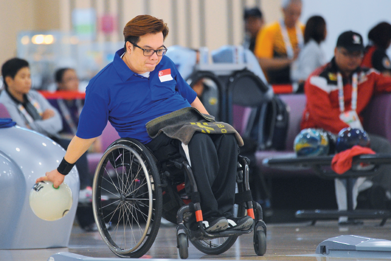 Imagem: Fotografia. Um homem com cabelos lisos, óculos, usando camisa azul e calça preta, sentado em uma cadeira de rodas. Ele segura com a mão esquerda uma bola de boliche. No fundo pessoas sentadas em cadeiras.  Fim da imagem.