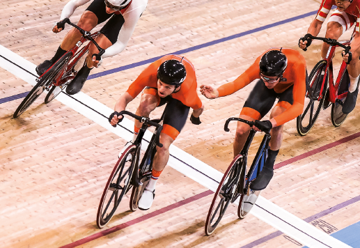 Imagem: Fotografia. Homens usando uniforme e capacetes sobre bicicletas em uma pista.  Fim da imagem.