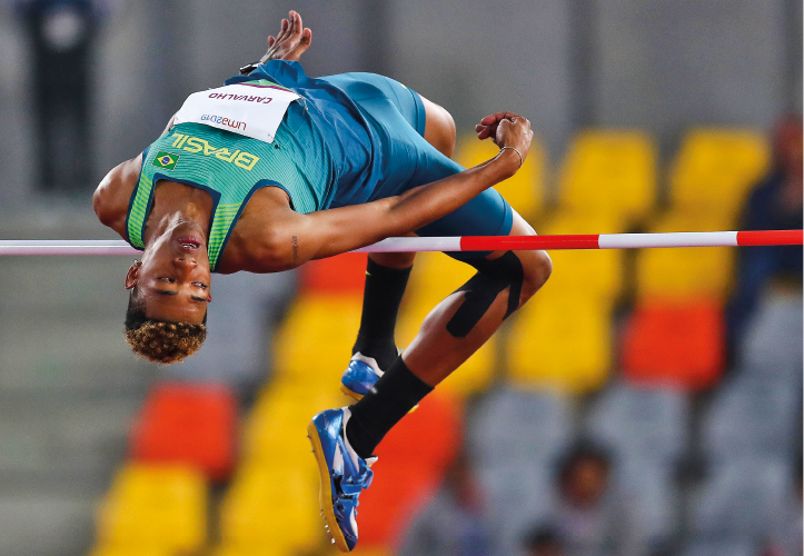 Imagem: Fotografia. Um homem negro, usando uniforme verde, com o tronco em formato em forma de arco sobre uma vara.  Fim da imagem.