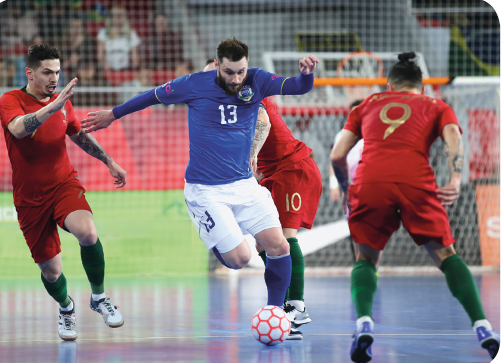 Imagem: Fotografia. Um homem com uniforme azul com uma bola próximo ao pé um uma quadra, ao redor dele, três homens com uniforme vermelho. Fim da imagem.