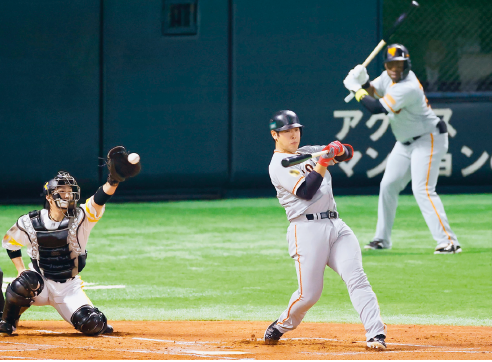 Imagem: Fotografia. À esquerda, um homem com capacete, luvas, proteção do tronco e pernas. Ele está com a mão esquerda esticada segurando uma bola de beisebol. Ao lado, um homem com uniforme branco e capacete, segurando um bastão na mão, sobre um campo marrom. Atrás, sobre o campo verde, outro homem usando uniforme branco, capacete e segurando um bastão.  Fim da imagem.