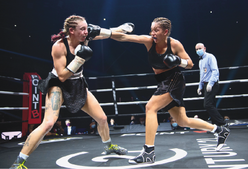 Imagem: Fotografia. Duas mulheres, usando luvas de boxes e roupas pretas dentro de um ringue. Elas estão uma de frente para a outra com o braço esticado na direção do rosto da outra. Atrás, um homem usando máscara olha na direção delas. Fim da imagem.