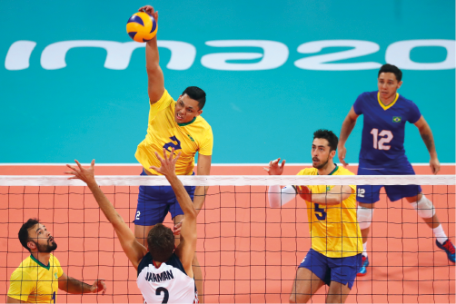 Imagem: Fotografia. Três homens usando camiseta amarela com números, um deles salta com a mão na bola que está no ar próximo à rede. De costas um homem com camiseta branca com número, com os braços esticados próximo a rede. Ao fundo, um homem de camiseta azul. Fim da imagem.