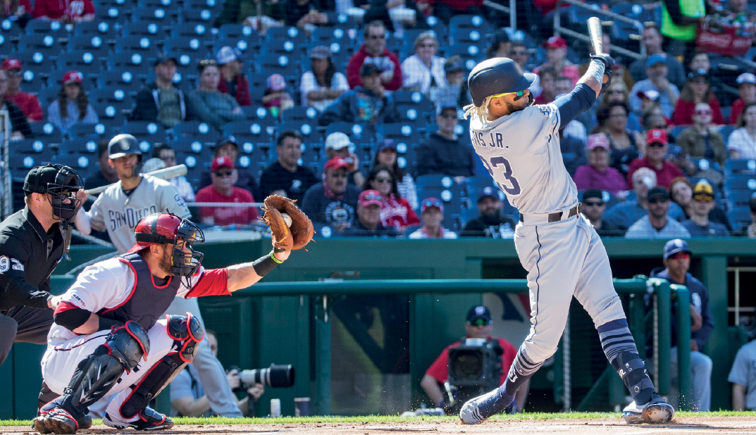 Imagem: Fotografia. À esquerda, com um capacete, o árbitro. Seguido de um homem com capacete, proteções e uma luva, Catcher ou receptor. Seguido de um homem em pé, com boné e uniforme com um bastão na mão. No fundo, outro jogador com bastão e arquibancada com pessoas olhando na direção deles.  Fim da imagem.