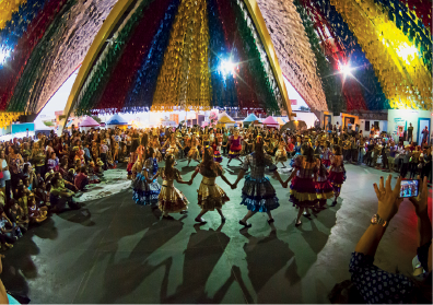 Imagem: Fotografia. Um grupo com roupas coloridas, de mãos dadas fazendo uma roda. Há ao redor diversas pessoas olhando para eles. Na parte superior, decoração com tiras de chita.  Fim da imagem.