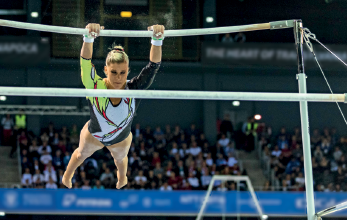 Imagem: Fotografia. Uma mulher com collant, com as mãos sobre uma barra. Na frente, outra barra.  Fim da imagem.