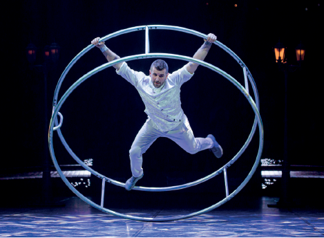 Imagem: Fotografia. Um homem com roupas brancas, segurando com as mãos e os pés um círculo gigante.  Fim da imagem.