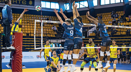 Imagem: Fotografia. À esquerda, um homem com uniforme amarelo. Ele salta, com o braço estendido na direção da bola, que está no ar, próximo à rede. Ao lado, quatro homens com uniforme amarelo olhando na direção da bola. À direita, três homens com uniforme verde. Eles saltam com os braços estendidos, próximo â rede, na direção da bola.  Fim da imagem.