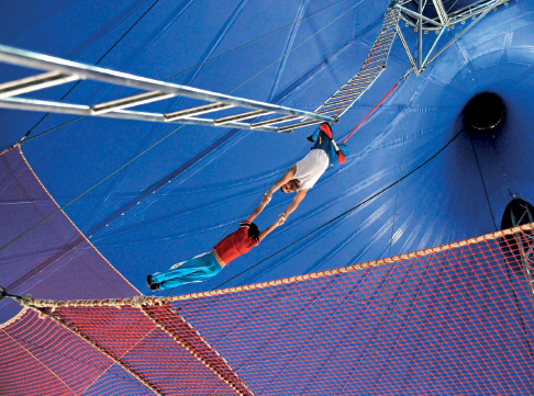 Imagem: Fotografia. Um homem de ponta cabeça, com as pernas presas em um trapézio, segurando os braços de uma pessoa que balança. Abaixo, uma rede de proteção.  Fim da imagem.