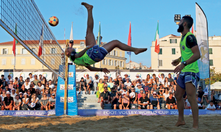 Imagem: Fotografia. Um homem de uniforme, ele está com as pernas no ar na direção de uma bola, próximo à rede, em um campo de areia. Ao lado, outro homem de uniforme, em pé olhando na direção da bola.  Fim da imagem.
