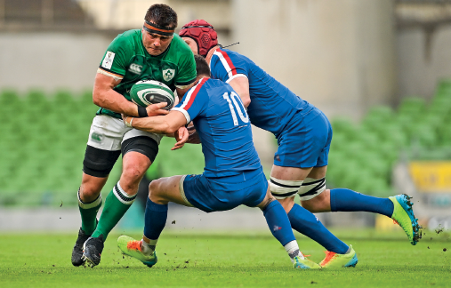 Imagem: Fotografia. Um homem com uniforme verde, segurando uma bola oval, ao redor dele, dois homens com uniforme azul, segura o corpo dele com as mãos em um campo com gramado verde. Fim da imagem.