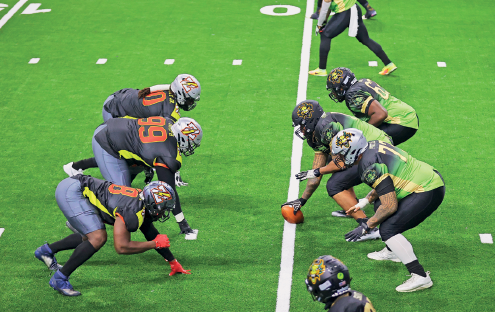 Imagem: Fotografia. À esquerda uma fileira com três homens de capacete e uniforme azul, ao lado, uma linha branca dividindo o campo. À direita, uma fileira de homens usando capacete e uniforme verde. Um deles está segurando uma bola oval em cima da linha branca. Fim da imagem.
