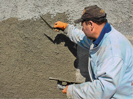 Imagem: Fotografia. Um homem de casaco azul e boné passando massa em uma parede.  Fim da imagem.