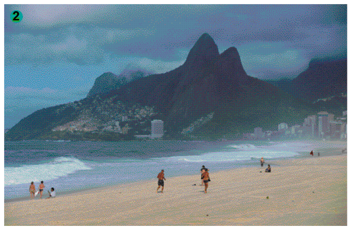 Imagem: Fotografia. Uma praia com algumas pessoas espalhadas. No fundo, prédios na beira da praia e morros com vegetação. O céu está nublado.   Fim da imagem.