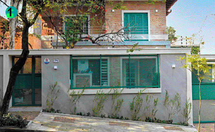 Imagem: Fotografia. Um sobrado de tijolos com varanda e uma grande janela de vidro no muro da frente.  Fim da imagem.