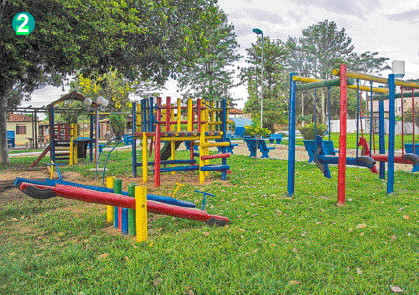 Imagem: Fotografia. Um parquinho feito de madeiras coloridas em uma área gramada. Há uma casinha, balanços e gangorras.  Fim da imagem.