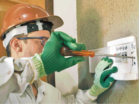 Imagem: Fotografia Um homem usando jaleco branco, capacete de proteção e luvas. Ele usa uma chave de fenda para mexer em uma caixa de energia na parede.  Fim da imagem.