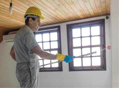 Imagem: Fotografia. Um homem de cabelo preto usando uma camiseta cinza e capacete de proteção amarelo. Ele pinta uma parede com um rolo. Na parede do fundo tem duas janelas.  Fim da imagem.