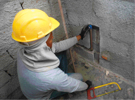Imagem: Fotografia. Um homem com camiseta cinza, pano na cabeça e capacete amarelo na cabeça. Ele coloca um cano em um buraco na parede.   Fim da imagem.
