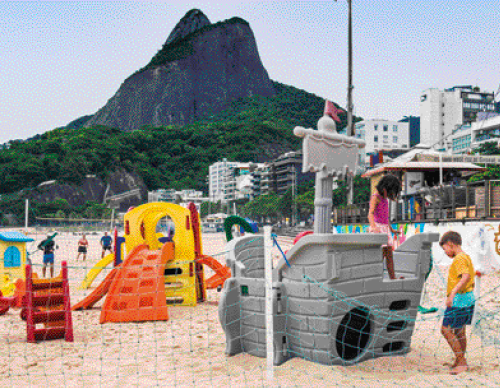 Imagem: Fotografia. Um parque com brinquedos de plástico na areia da praia, cercado por uma rede verde. Tem um brinquedo em forma de navio cinza, com uma menina de cabelo preto e roupa rosa em cima e um menino loiro de camiseta amarela do lado. Atrás, uma casinha e uma torre com escorregados. No fundo da praia tem pessoas jogando vôlei na areia, prédios e um morro.  Fim da imagem.