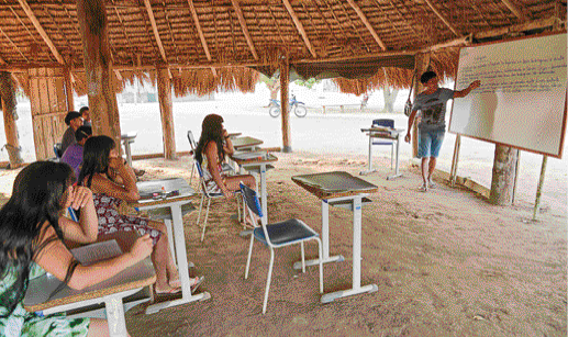 Imagem: Fotografia. Uma sala de aula embaixo de uma cobertura de palha, sem fechamento lateral. De um dos lados, uma mulher aponta para uma lousa branca que está em um suporta. Na frente dela, mesas e cadeiras enfileiradas com alguns alunos sentados.  Fim da imagem.