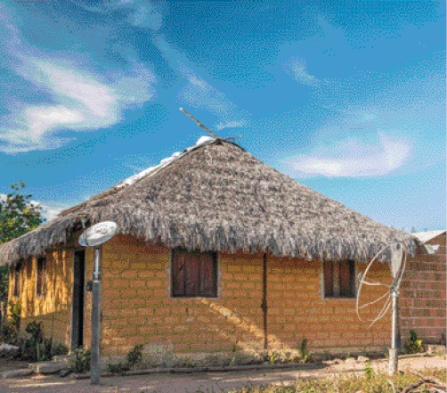 Imagem: Fotografia. Uma casa com paredes de tijolo, janelas de madeira e telhado de palha. Na frente tem uma antena.  Fim da imagem.