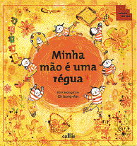 Imagem: Ilustração. Capa de livro. No centro, um círculo com o título: minha mão é uma régua. Ao redor há crianças brincando, flores e brinquedos.   Fim da imagem.