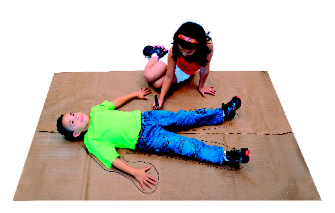 Imagem: Fotografia. Um menino de cabelo preto usando camiseta verde, calça jeans e um tênis. Ele está deitado em cima de folhas de papel pardo, com os braços e pernas abertas. Ao lado, uma menina de cabelo preto e liso usando uma saia branca e camiseta laranja está sentada usando uma caneta para fazer o contorno do corpo do menino na folha.  Fim da imagem.