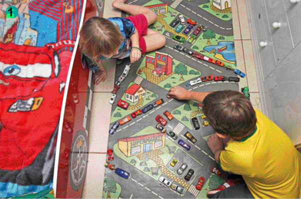 Imagem: Fotografia. Uma menina loira usando uma bermuda vermelha e camiseta azul e um menino loiro de cabelo liso e camiseta amarela. Eles estão sentados em cima de um tapete com o desenho de uma cidade com ruas e há carrinhos de brinquedo nas ruas.   Fim da imagem.