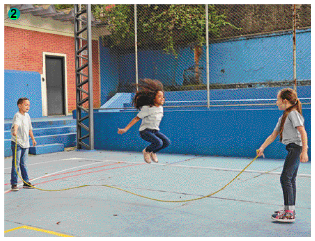 Imagem: Fotografia. Uma menina negra de calça jeans e camiseta branca pulando corda que está sendo segurada por duas outras crianças: um menino de cabelo calça jeans e camiseta branca e uma menina ruiva de rabo de cavalo com calça jeans e camiseta branca. Eles estão em uma quadra de esportes.   Fim da imagem.