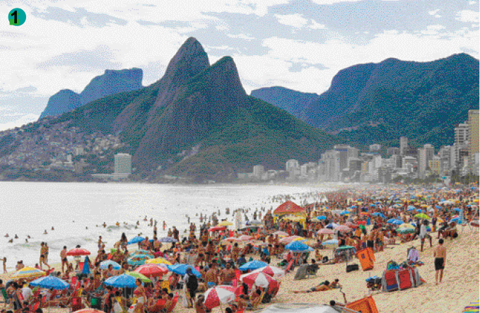 Imagem: Fotografia. Uma praia lotada com vários guarda-sóis coloridos na areia, com pessoas entre eles. No fundo tem alguns prédios ao lado da praia e morros com vegetação. O céu está azul.  Fim da imagem.