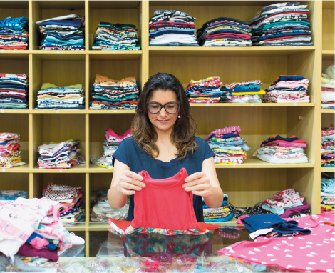 Imagem: Fotografia. Uma mulher de cabelo castanho usando uma camiseta azul. Ela segura uma peça de roupa vermelha nas mãos. Atrás dela, prateleiras com roupas de diversas cores dobradas. Fim da imagem.