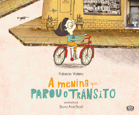 Imagem: Ilustração. Uma menina de cabelo preto e arco, óculos, camiseta azul e calça amarela. Ela está sorrindo de olhos fechados e anda de bicicleta. Atrás dela tem uma loja. Na parte de baixo, título do livro: a menina que parou o trânsito.  Fim da imagem.