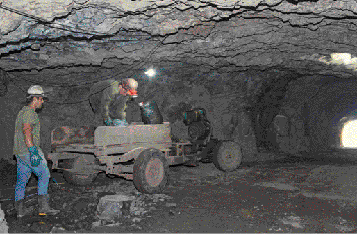 Imagem: Fotografia. Dois homens de camiseta verde e boné, um está de pé e o outro abaixado em um carrinho de metal. Eles estão em um túnel de terra.   Fim da imagem.