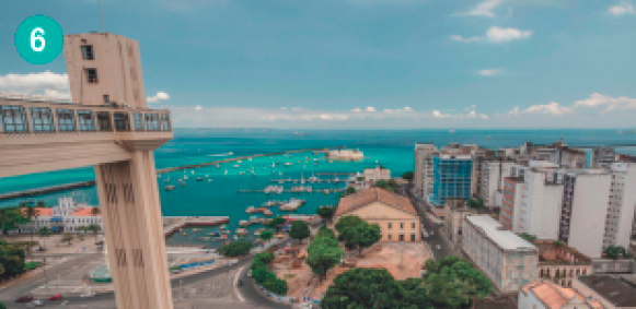 Imagem: Fotografia. Vista aérea de uma cidade na beira do mar. No canto esquerdo, uma torre com uma passarela.   Fim da imagem.