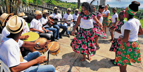 Imagem: Fotografia. Mulheres de blusa branca com golas florias e saias florias. Elas dançam na frente de homens que estão sentados enfileirados em cadeiras. Os homens tocam instrumentos musicais.   Fim da imagem.