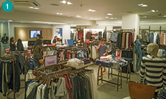 Imagem: Fotografia. Uma loja com araras e cabides com roupas coloridas e algumas pessoas olhando.   Fim da imagem.