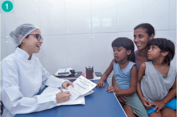 Imagem: Fotografia. Uma mulher morena com duas crianças no colo: uma menina de cabelo preto e liso com franja e vestido azul e um menino de cabelo preto e liso com uma regata cinza. Na frente deles há uma mesa azul. Do outro lado da mesa, uma mulher de jaleco branco e touca sorri. Ela segura papéis nas mãos.  Fim da imagem.