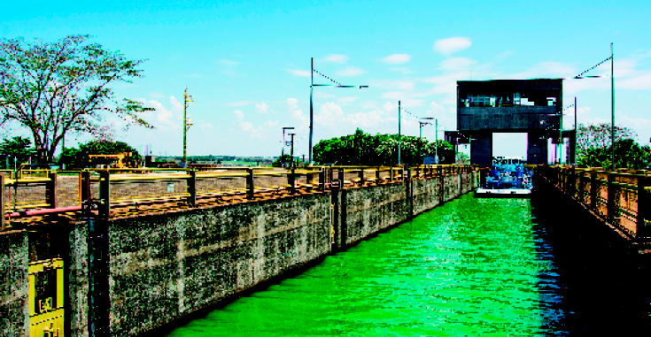 Imagem: Fotografia. Um barco passando por um canal margeado por duas avenidas.  Fim da imagem.