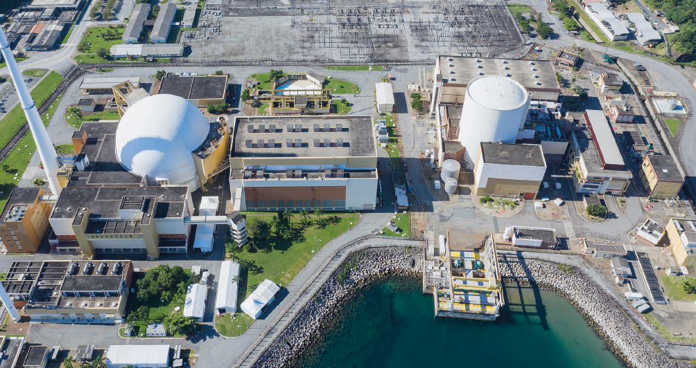 Imagem: Fotografia. Uma usina na beira da água. As construções são retangulares com uma semiesfera de um dos lados e uma cilíndrica do outro lado. Em um dos cantos, uma torre branca.  Fim da imagem.