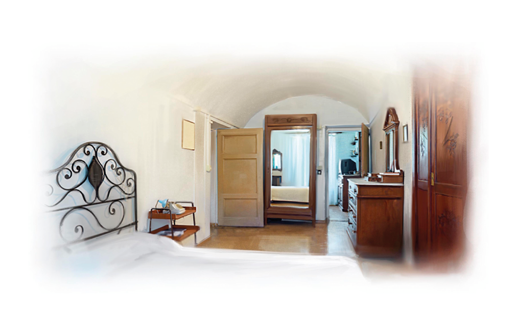 Imagem: Fotografia. Um quarto branco com cama de casal arrumada com cabeceira com arabescos e criado-mudo onde está um telefone antigo. Na outra parede estão uma penteadeira de madeira e um guarda-roupa. Ao fundo, um outro móvel de madeira com amplo espelho e uma porta que leva a outro cômodo.  Fim da imagem.