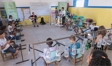 Imagem: Fotografia. Destaque do interior de uma sala onde as carteiras estão posicionas em semicírculo. As crianças estão sentadas diante de instrumentos e observam uma mulher tocar violão ao centro. Ao fundo, há garrafas vazias penduradas em um suporte de ferro. Na parede, há diversas colagens.  Fim da imagem.