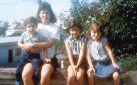 Imagem: Fotografia. Uma mulher sorri com uma criança pequena sentada em seu colo e ao seu lado estão duas meninas maiores. Todos têm pele clara, cabelo liso e sorriem. Estão em um banco próximo a arbustos com algumas casas ao fundo. Fim da imagem.