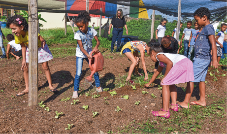 Imagem: Fotografia. Um grupo de crianças está acompanhado de duas mulheres adultas em uma horta coberta por uma rede. Algumas observam e outras regam as mudas.  Fim da imagem.